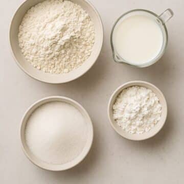 Top-down view of flour, sugar, cornstarch, and milk in ceramic bowls and glass, common ingredients replaced in keto dessert recipes.