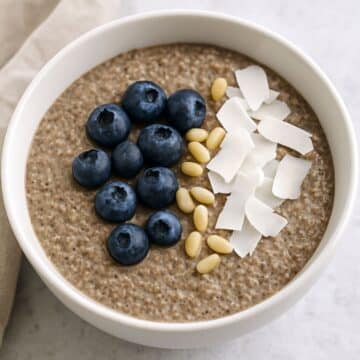Bowl of keto-friendly chia seed oatmeal with blueberries, coconut flakes, and pine nuts on a neutral background.